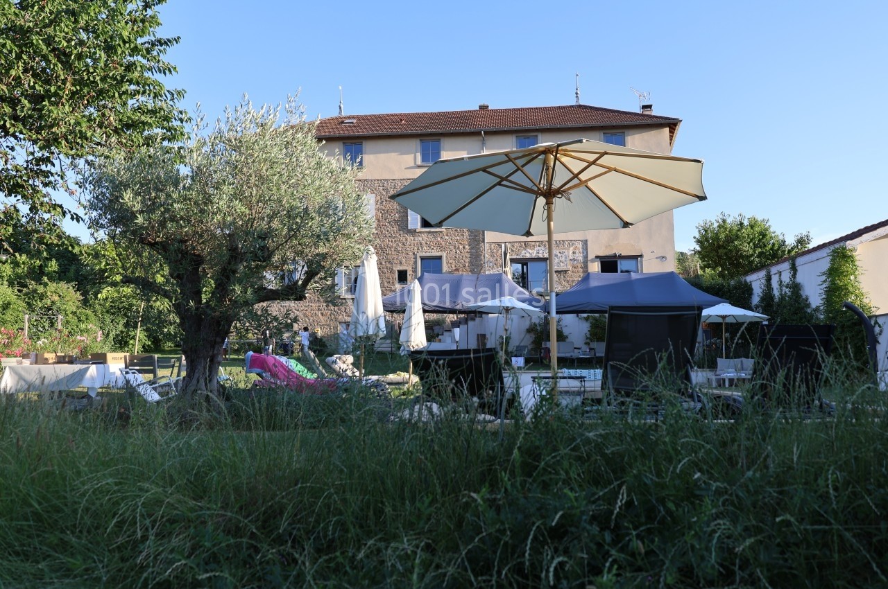 Jardin avec parasols, mobilier d'extérieur et un grand arbre, devant une maison en pierre à plusieurs étages.