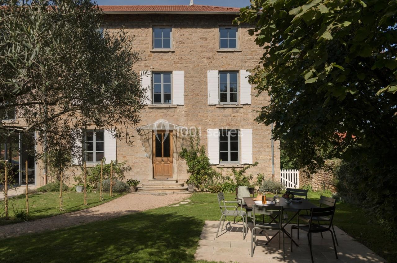 Façade en pierre d'une maison avec volets blancs, entourée d'un jardin arboré et d'une table extérieure dressée.