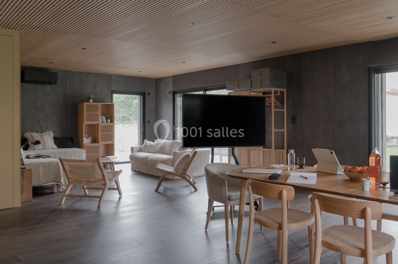 Salle de séjour moderne avec mobilier en bois, canapé clair, table de travail et grande télévision sur pied.