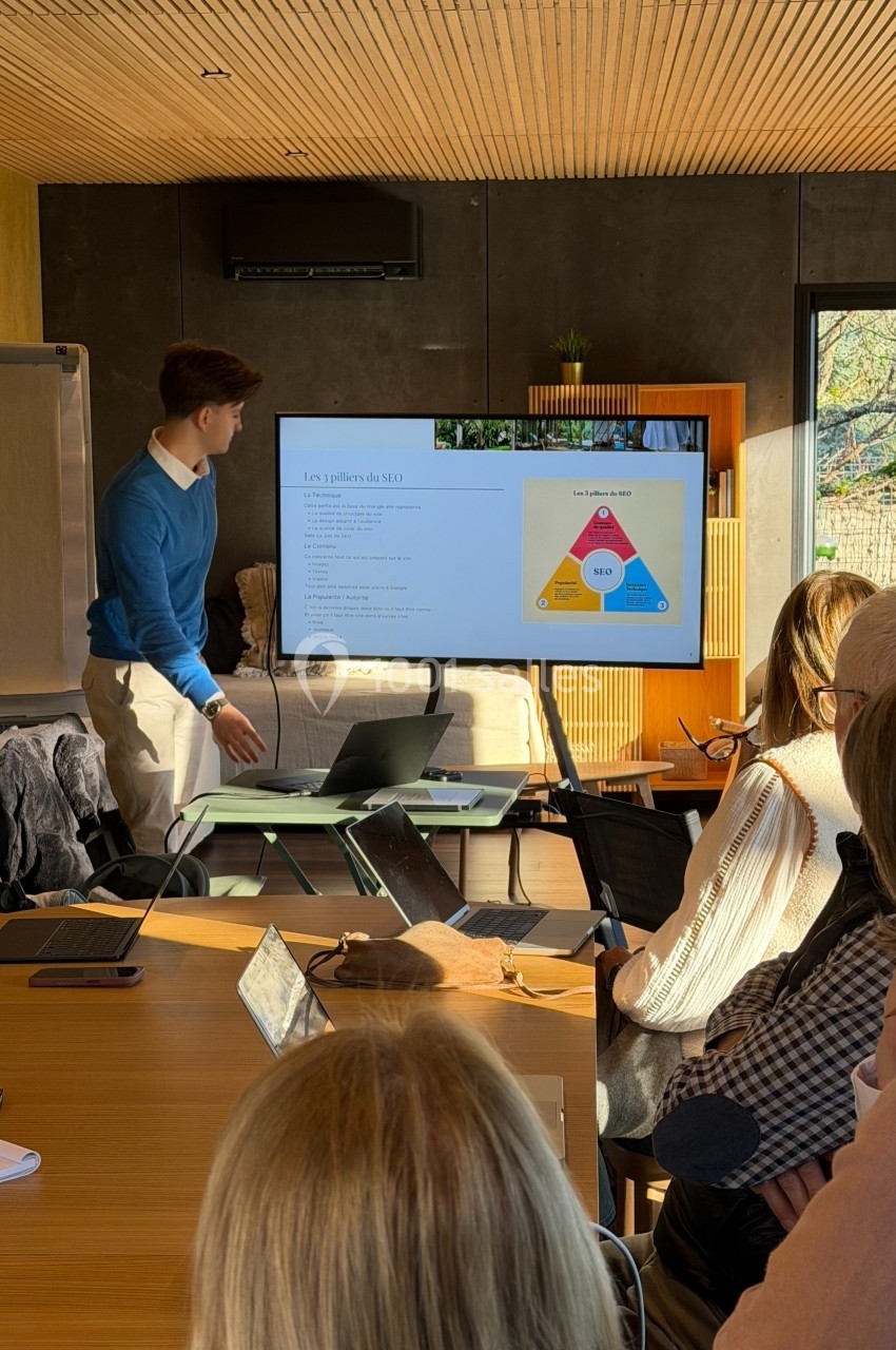 Un homme présente un diaporama sur un écran devant un groupe de personnes dans une salle lumineuse.