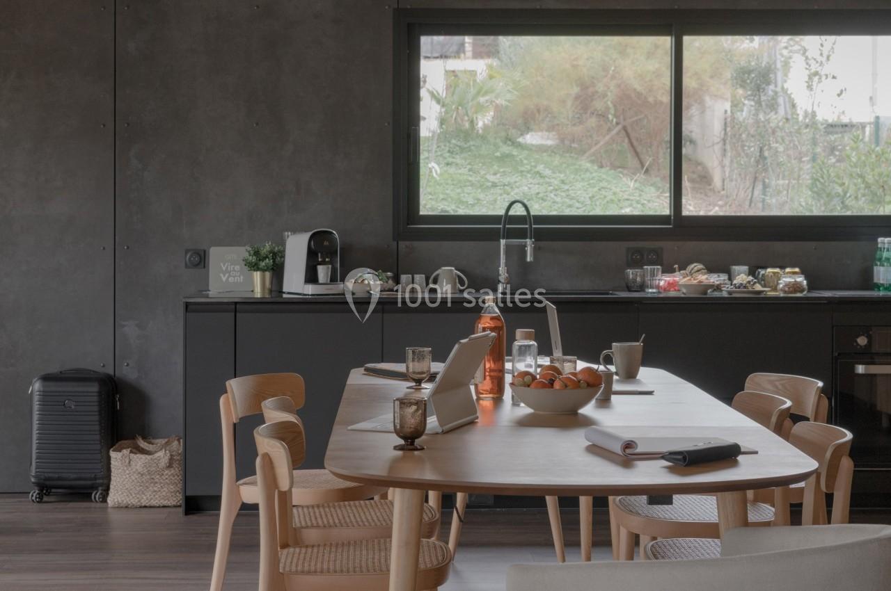 Salle à manger moderne avec table en bois, chaises assorties, cuisine ouverte et grande fenêtre donnant sur un jardin.