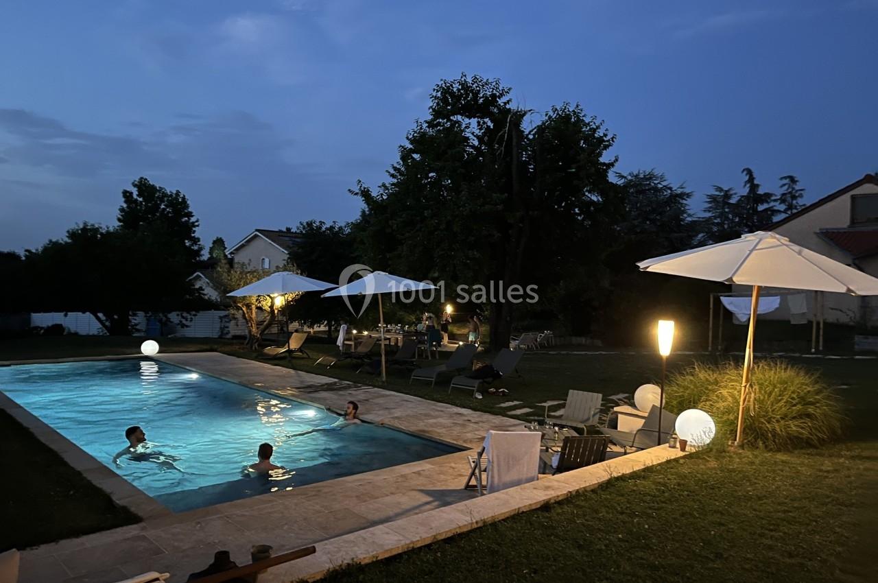 Piscine éclairée en soirée avec des personnes nageant, entourée de parasols, chaises longues et végétation.
