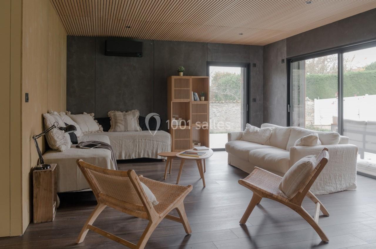 Salon moderne avec canapé blanc, fauteuils en bois, étagère, et grandes baies vitrées donnant sur un jardin.
