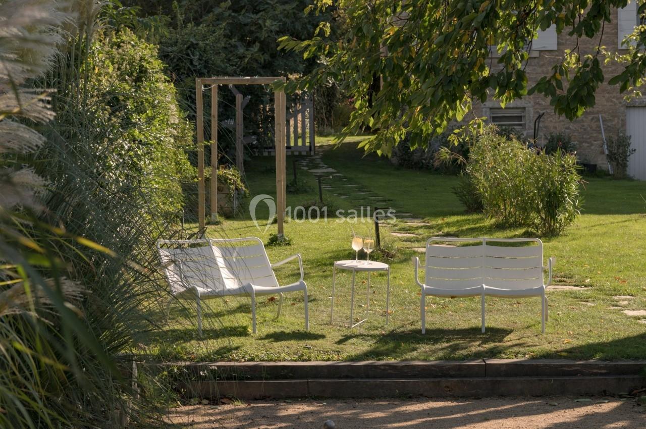 Deux chaises blanches et des verres de vin sur une table dans un jardin, avec un terrain de pétanque au premier plan.