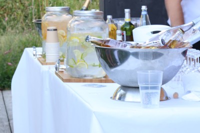 Table dressée en extérieur avec boissons fraîches, carafes d'eau infusée, bouteilles et verres sur une nappe blanche.