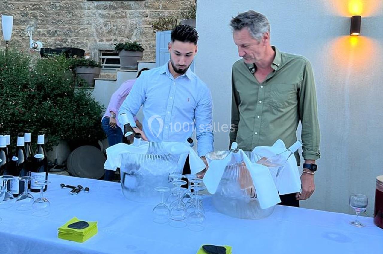 Deux hommes préparent des bouteilles de vin dans des seaux à glace sur une table dressée en extérieur.