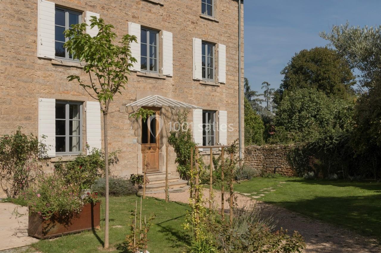 Façade en pierre d'une maison à trois étages avec volets blancs, jardin arboré et allée gravillonnée au premier plan.