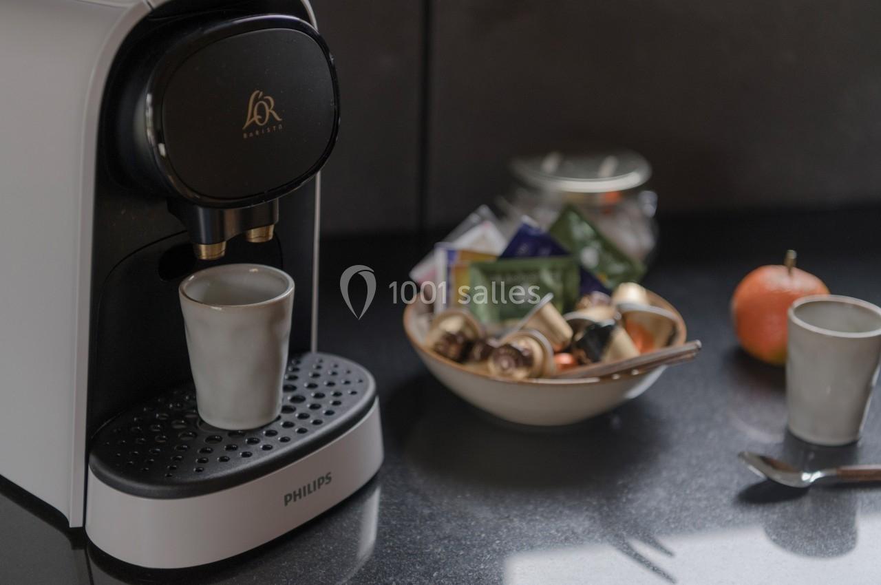 Machine à café avec une tasse en céramique, posée sur un plan de travail avec des capsules et une orange en arrière-plan.