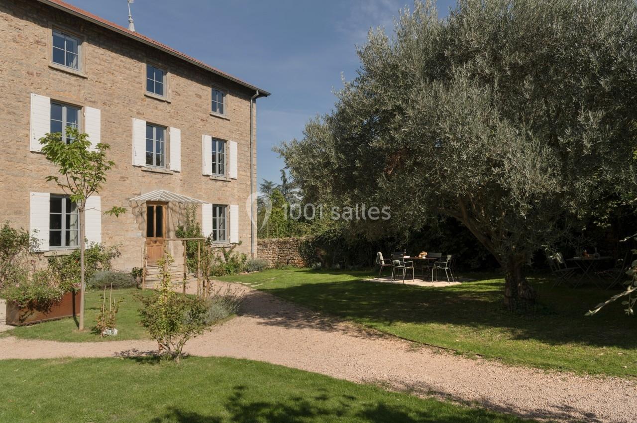 Maison en pierre avec volets blancs entourée d'un jardin arboré, table et chaises installées sous un olivier.