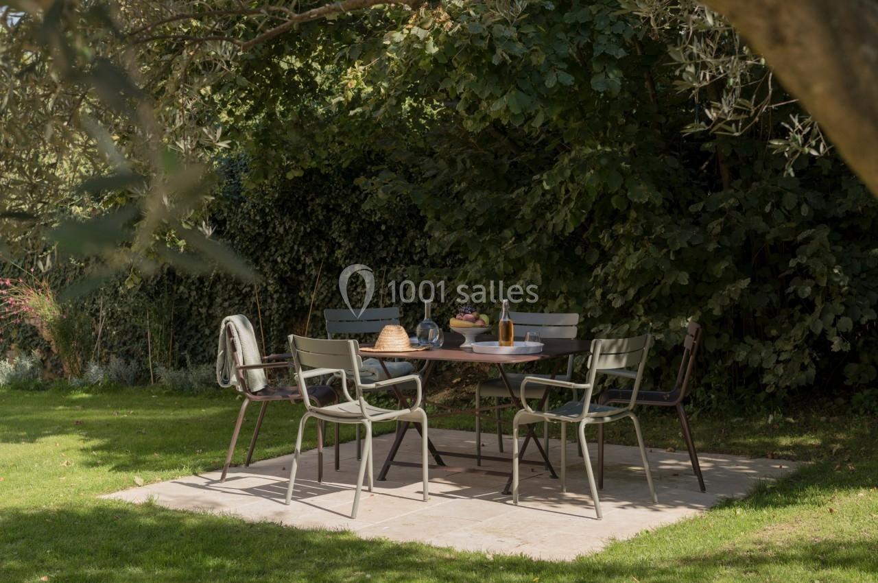 Table de jardin entourée de chaises sur une terrasse en pierre, ombragée par des arbres et entourée de verdure.