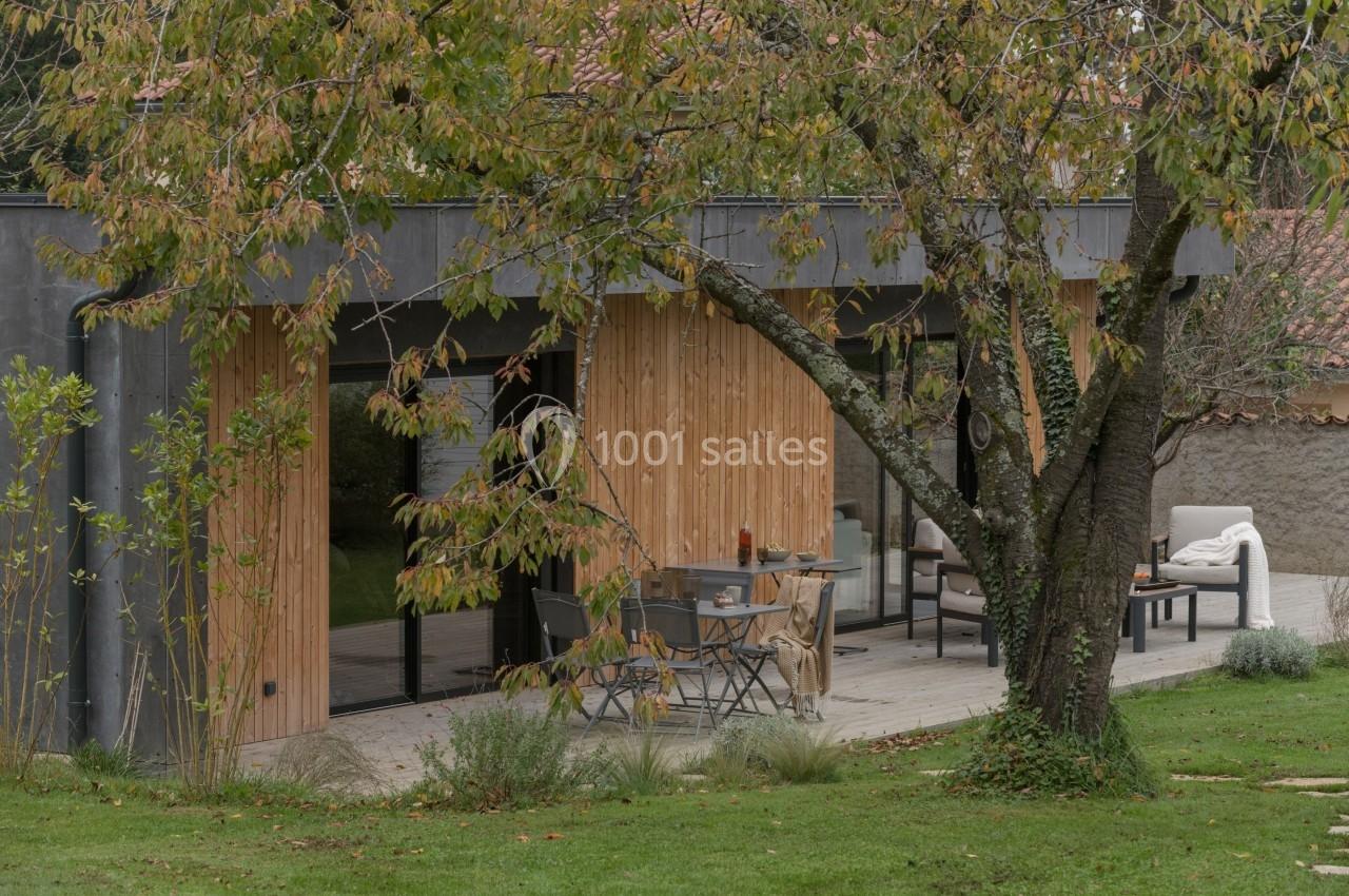 Terrasse en bois aménagée avec table et chaises, adossée à une maison moderne en bois, entourée de verdure.