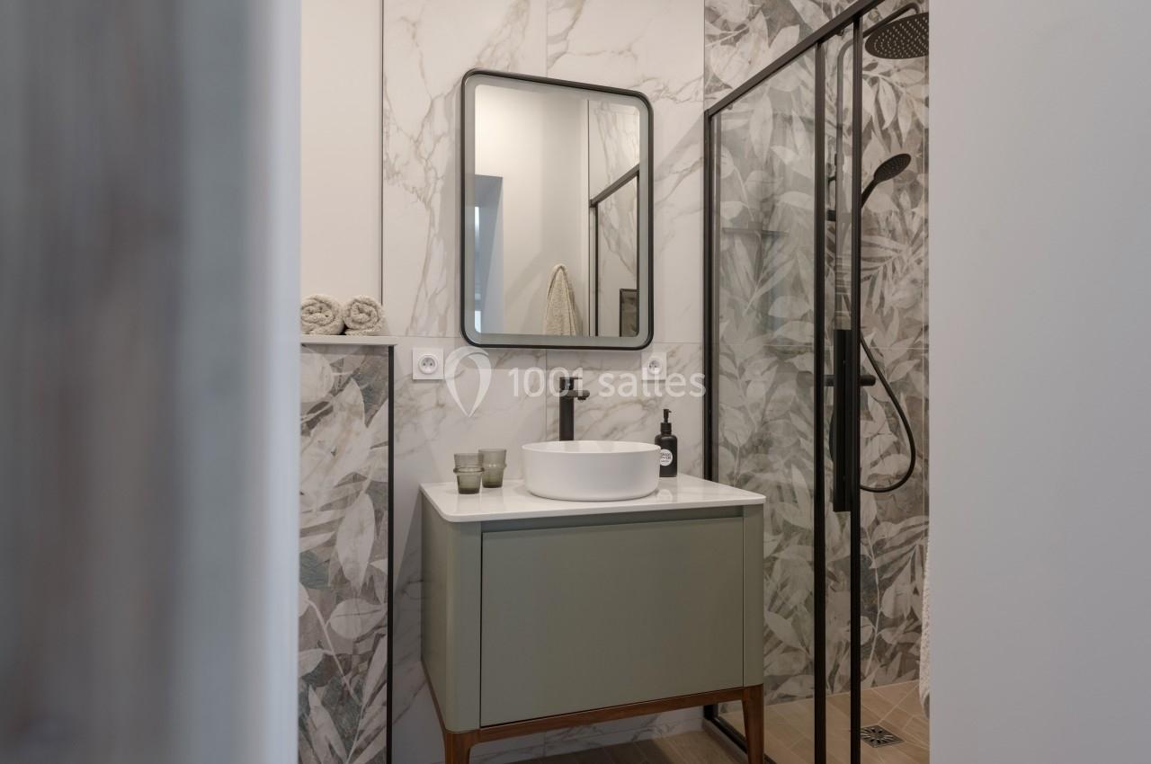 Salle de bain moderne avec lavabo sur meuble vert, miroir rectangulaire et douche à paroi vitrée avec motifs végétaux.