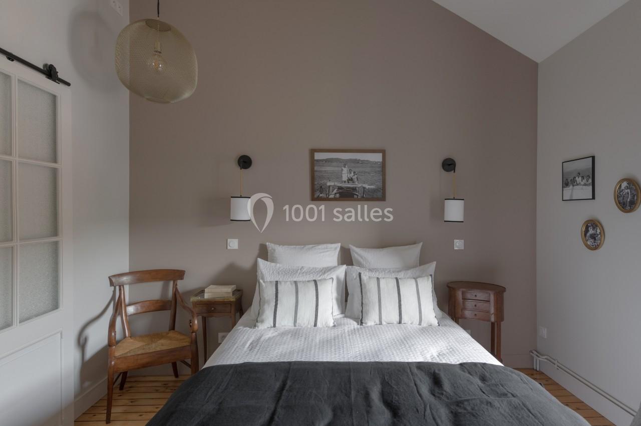 Chambre avec lit double, murs taupe, décor sobre, suspension en rotin, et mobilier en bois.