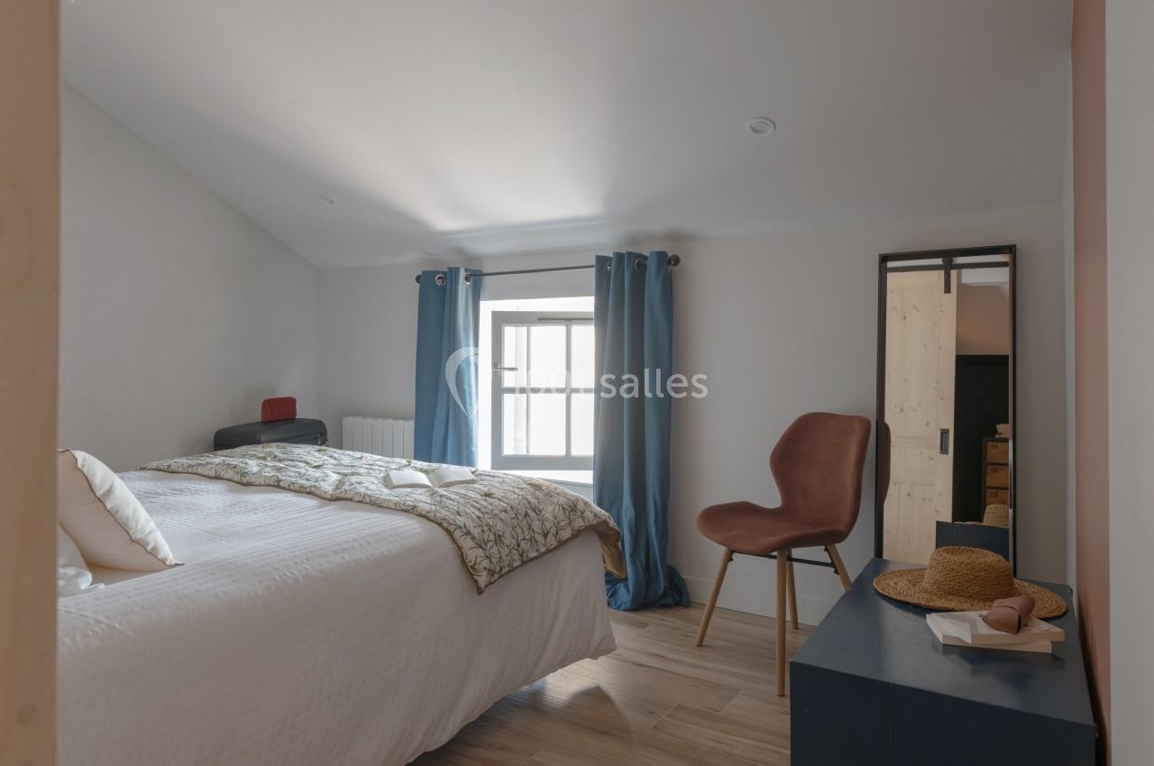 Chambre lumineuse avec lit double, chaise rose, miroir, rideaux bleus et décoration simple sur une commode.