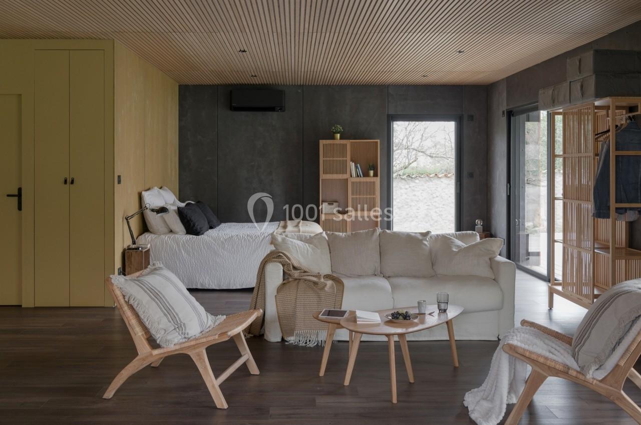 Salon ouvert avec canapé beige, fauteuils en bois, table basse, lit en arrière-plan et grandes fenêtres laissant entrer la…