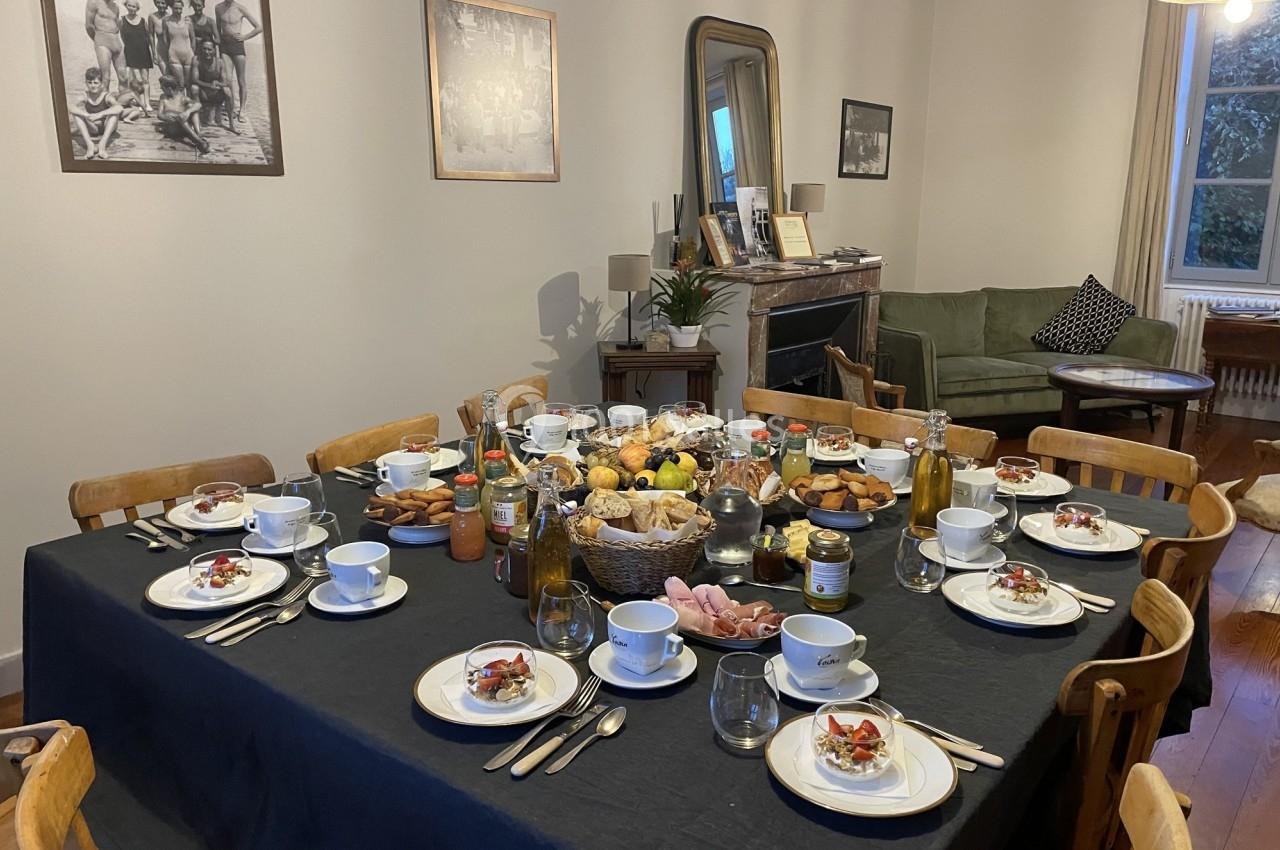 Table dressée pour un repas, avec assiettes, couverts, bols, et divers plats au centre dans une salle à manger lumineuse.