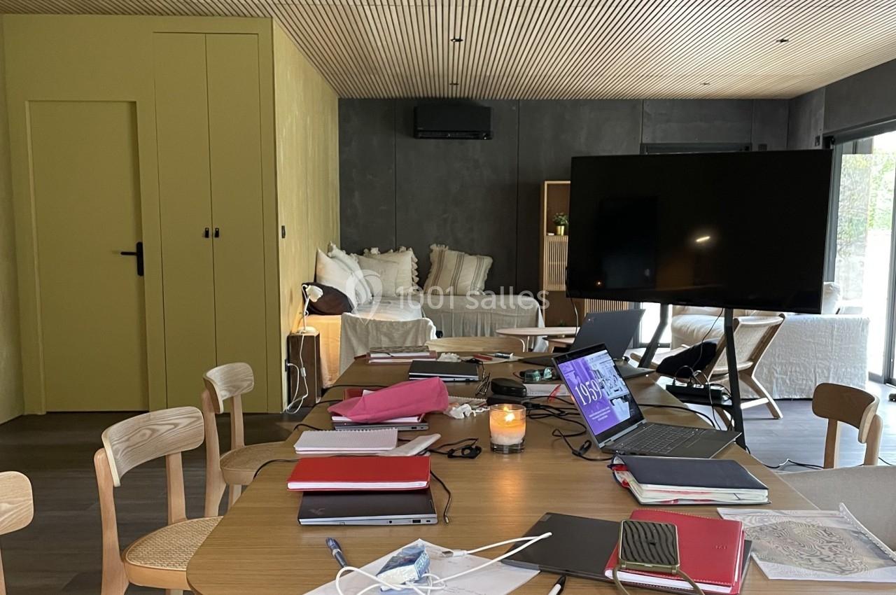 Salle de réunion moderne avec table encombrée de documents, chaises en bois, canapé et grand écran en arrière-plan.