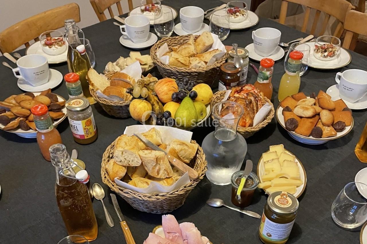 Table dressée pour un petit-déjeuner avec pains, fruits, confitures, viennoiseries, fromages et boissons variées.