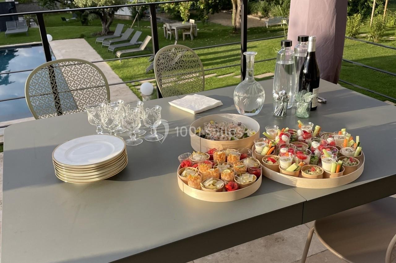 Table dressée en extérieur avec verrines colorées, assiettes empilées, verres, carafe et bouteilles, jardin en arrière-plan.