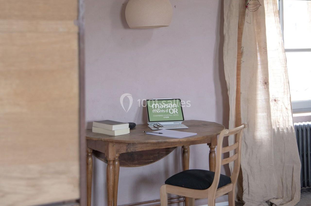 Bureau en bois avec ordinateur portable allumé, cahier et stylo, près d'une fenêtre avec rideaux beiges.