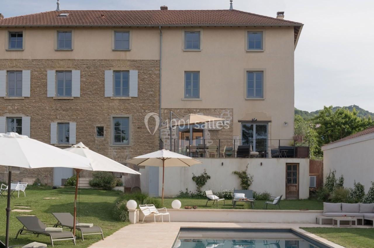 Façade d'une maison en pierre et crépi avec jardin, terrasse, piscine et mobilier extérieur sous des parasols.