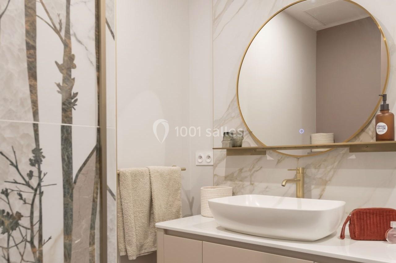 Salle de bain moderne avec vasque blanche, miroir rond doré, mur en marbre et paroi de douche décorée de motifs végétaux.