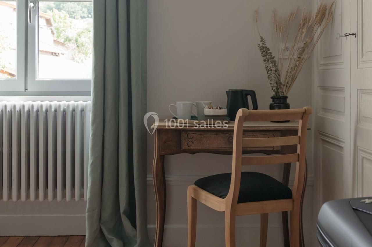 Petit bureau en bois avec une chaise, une cafetière, des tasses et des plantes séchées, près d'une fenêtre avec rideaux…