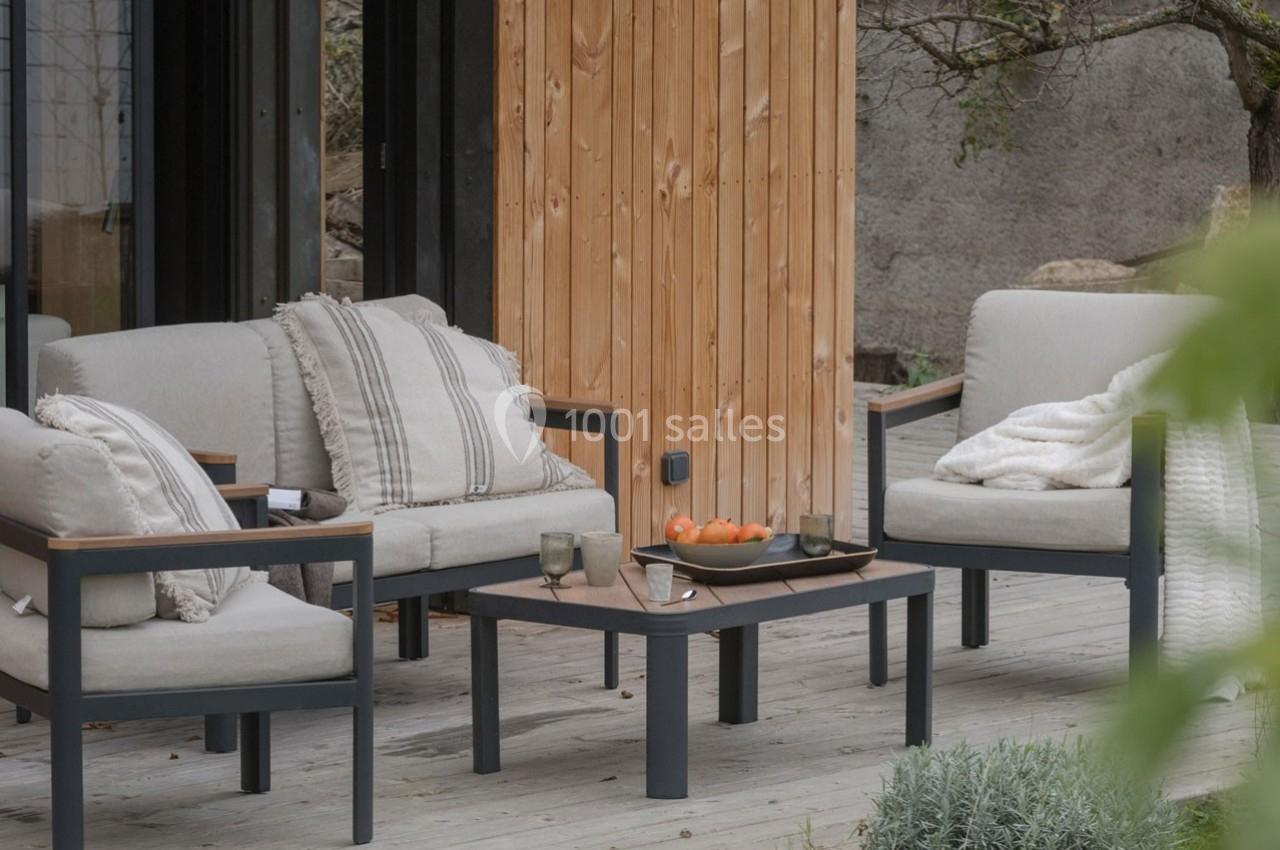 Mobilier de jardin avec coussins beiges, table basse et bol de fruits sur une terrasse en bois près d'un mur en bois clair.