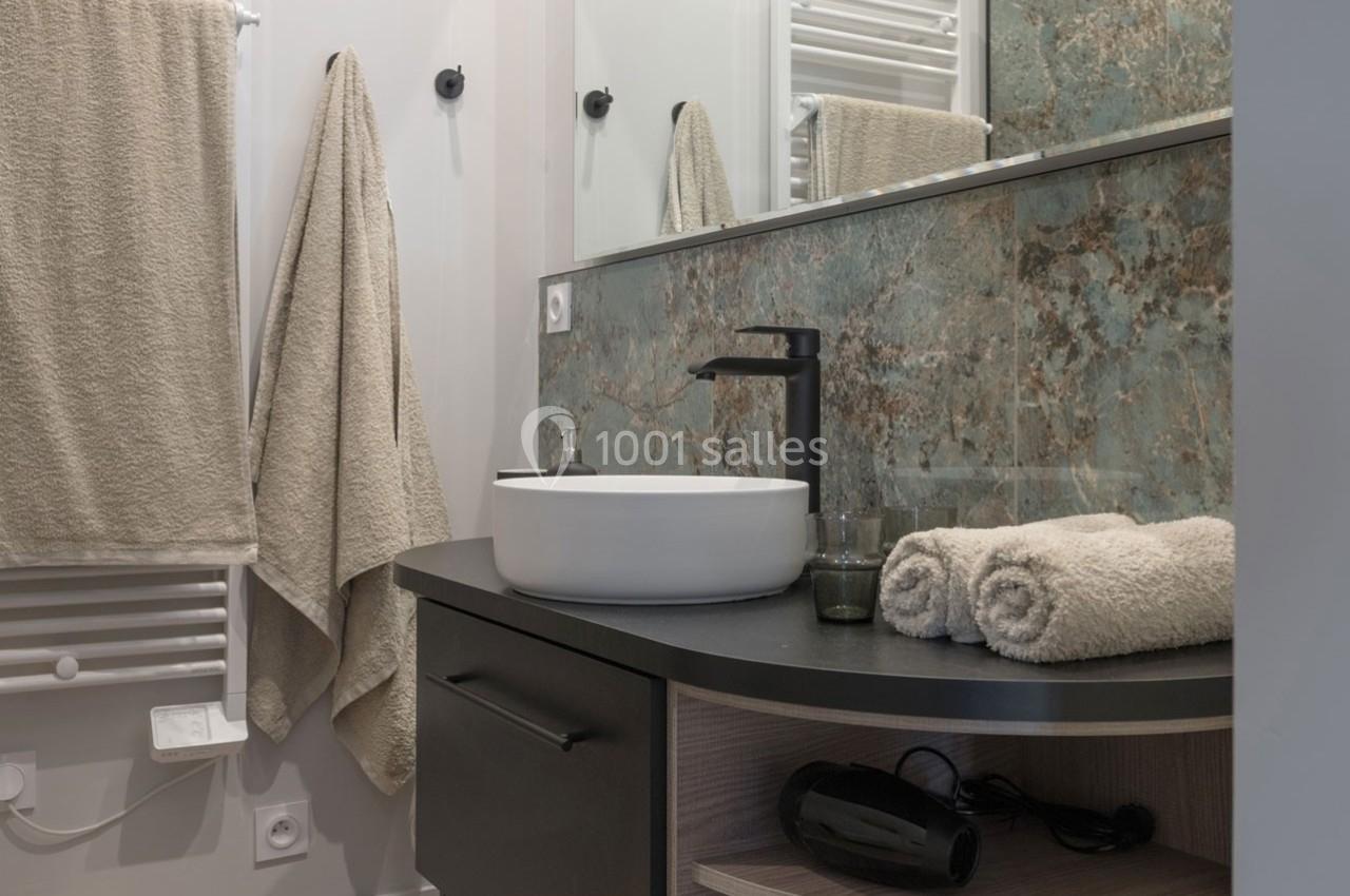 Salle de bain moderne avec lavabo blanc, robinet noir, miroir, serviettes beiges et sèche-cheveux sur une étagère.
