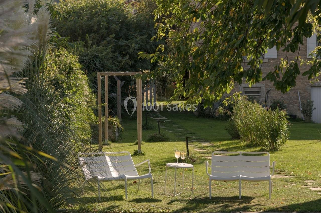 Deux chaises blanches et une table avec des verres de vin dans un jardin verdoyant, sous des arbres.