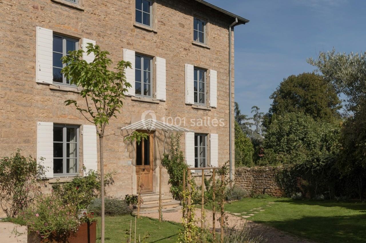 Façade d'une maison en pierre avec volets blancs, jardin arboré et allée ensoleillée au premier plan.