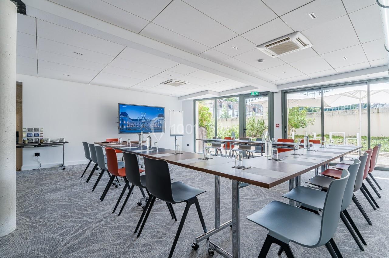 Salle de réunion lumineuse avec grandes baies vitrées, tables disposées en U et écran de présentation.