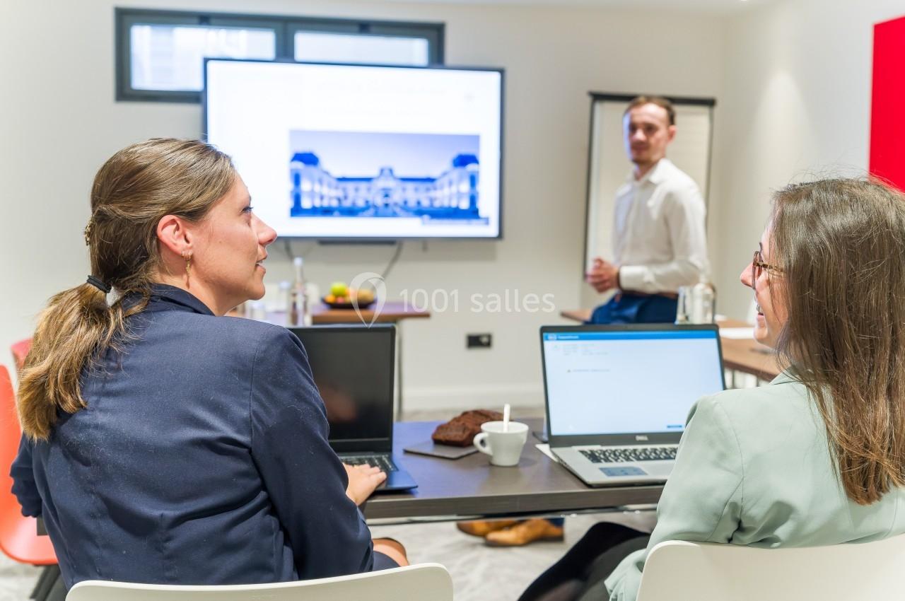 Deux femmes travaillent sur des ordinateurs portables dans une salle de réunion, un homme présente devant un écran.