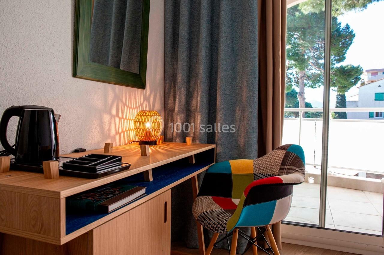 Bureau en bois avec lampe allumée, chaise colorée et vue sur un balcon donnant sur des arbres et des bâtiments.