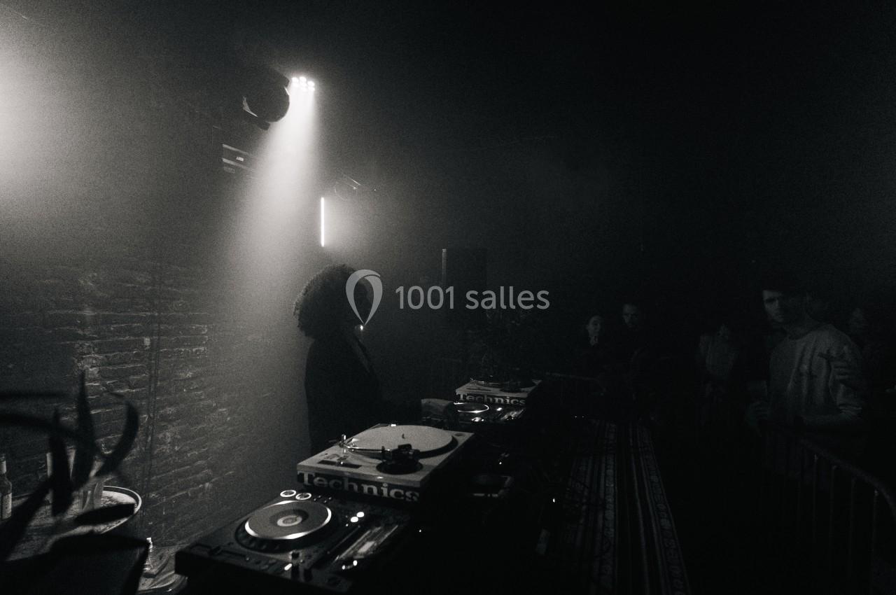 Une DJ mixe sur des platines dans une ambiance sombre, face à un public dans une salle aux murs en briques.