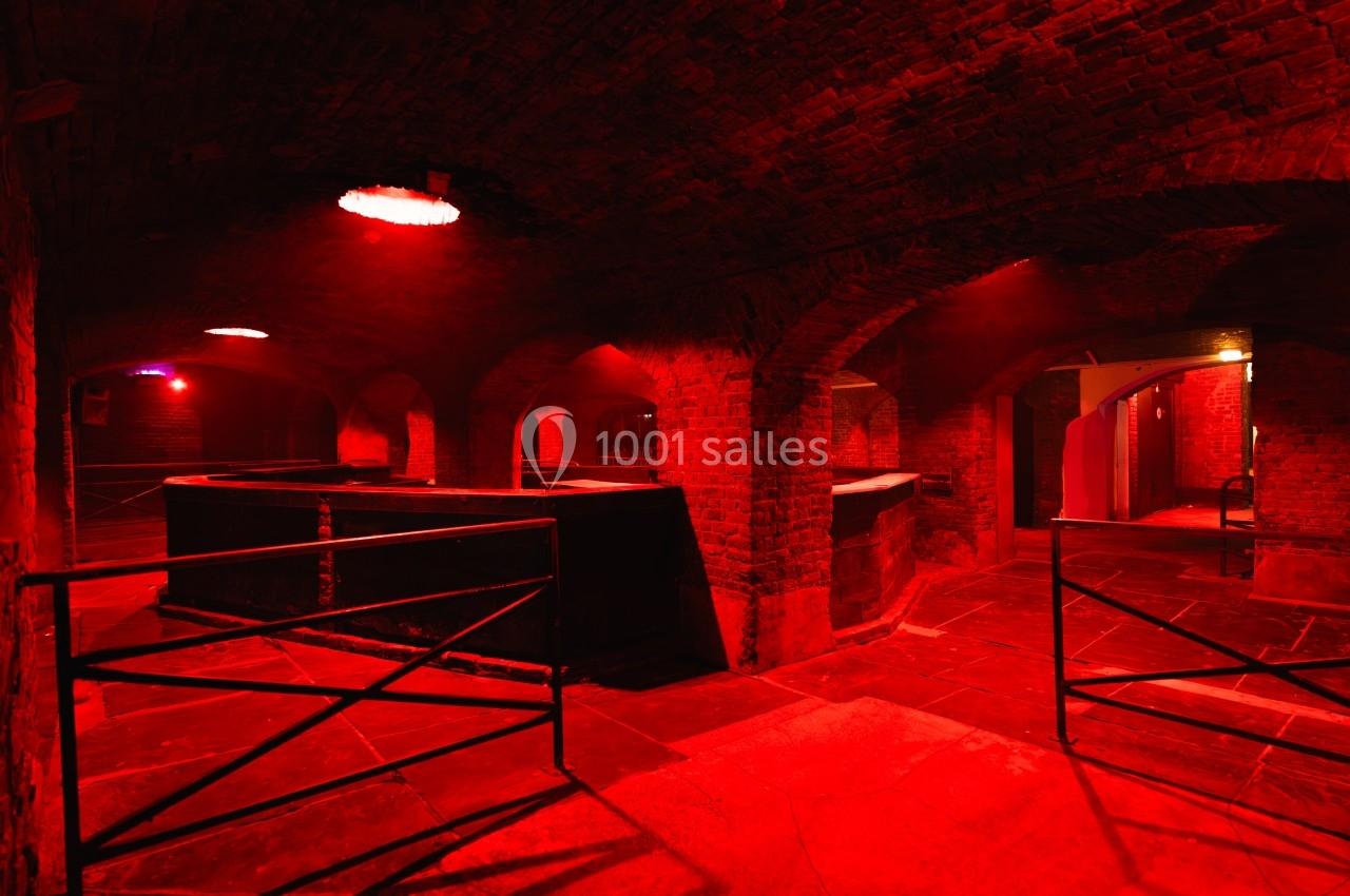 Salle voûtée en briques éclairée par une lumière rouge, avec des barrières métalliques et un sol en pierre.