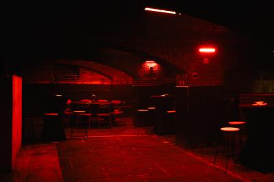 Salle sombre avec murs en briques, éclairage tamisé, scène en bois et tables basses au premier plan.