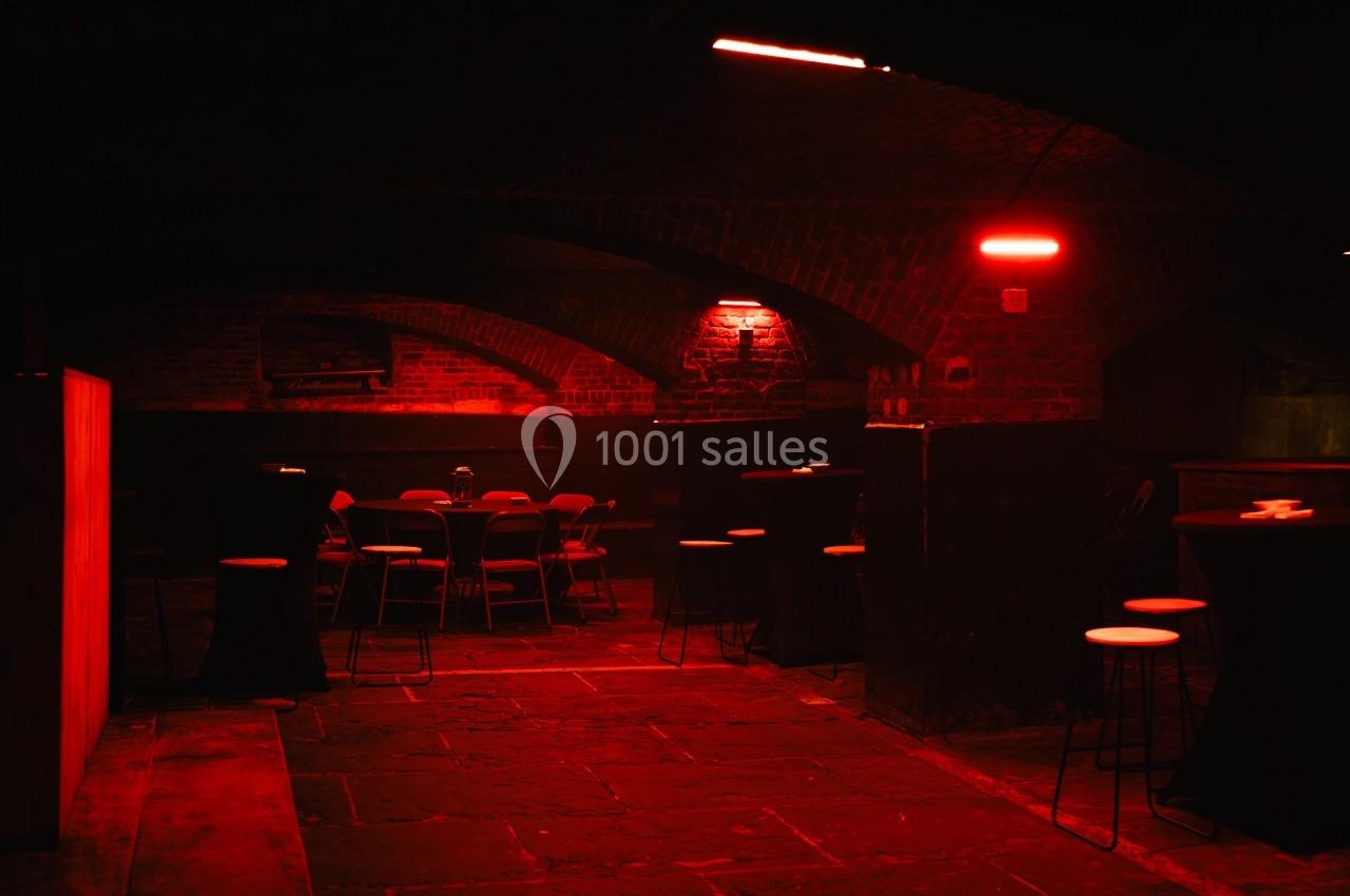 Salle voûtée en briques éclairée par une lumière rouge, avec tables hautes et chaises dispersées.