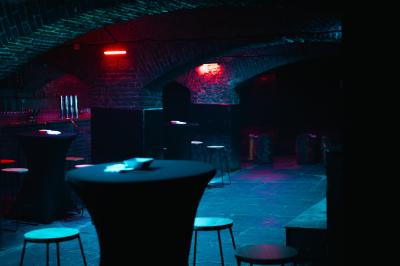 Salle sombre avec murs en briques, éclairage tamisé, scène en bois et tables basses au premier plan.