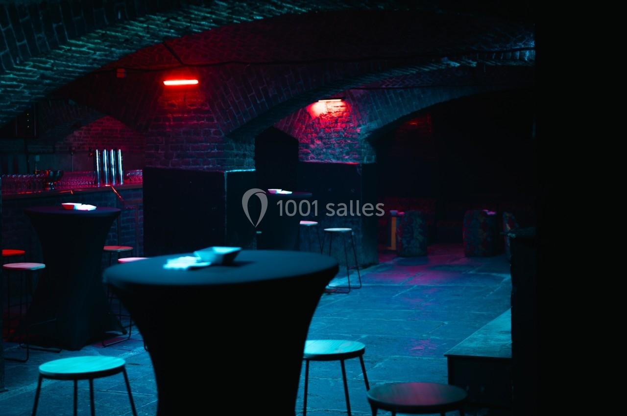 Salle voûtée sombre avec éclairage rouge et bleu, tables hautes noires et tabourets dispersés.