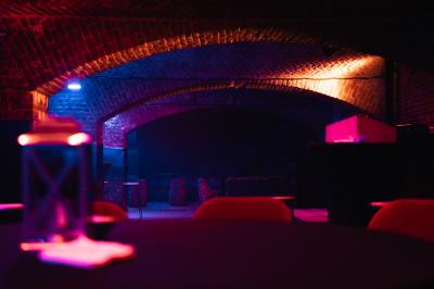 Salle sombre avec murs en briques, éclairage tamisé, scène en bois et tables basses au premier plan.