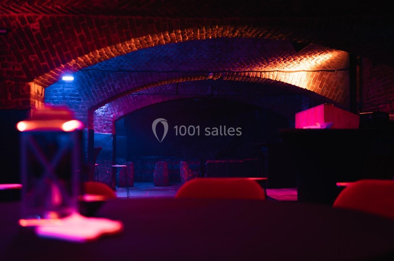 Salle voûtée en briques avec éclairage tamisé coloré, tables et chaises visibles au premier plan.