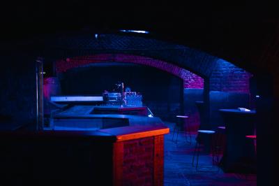 Salle sombre avec murs en briques, éclairage tamisé, scène en bois et tables basses au premier plan.