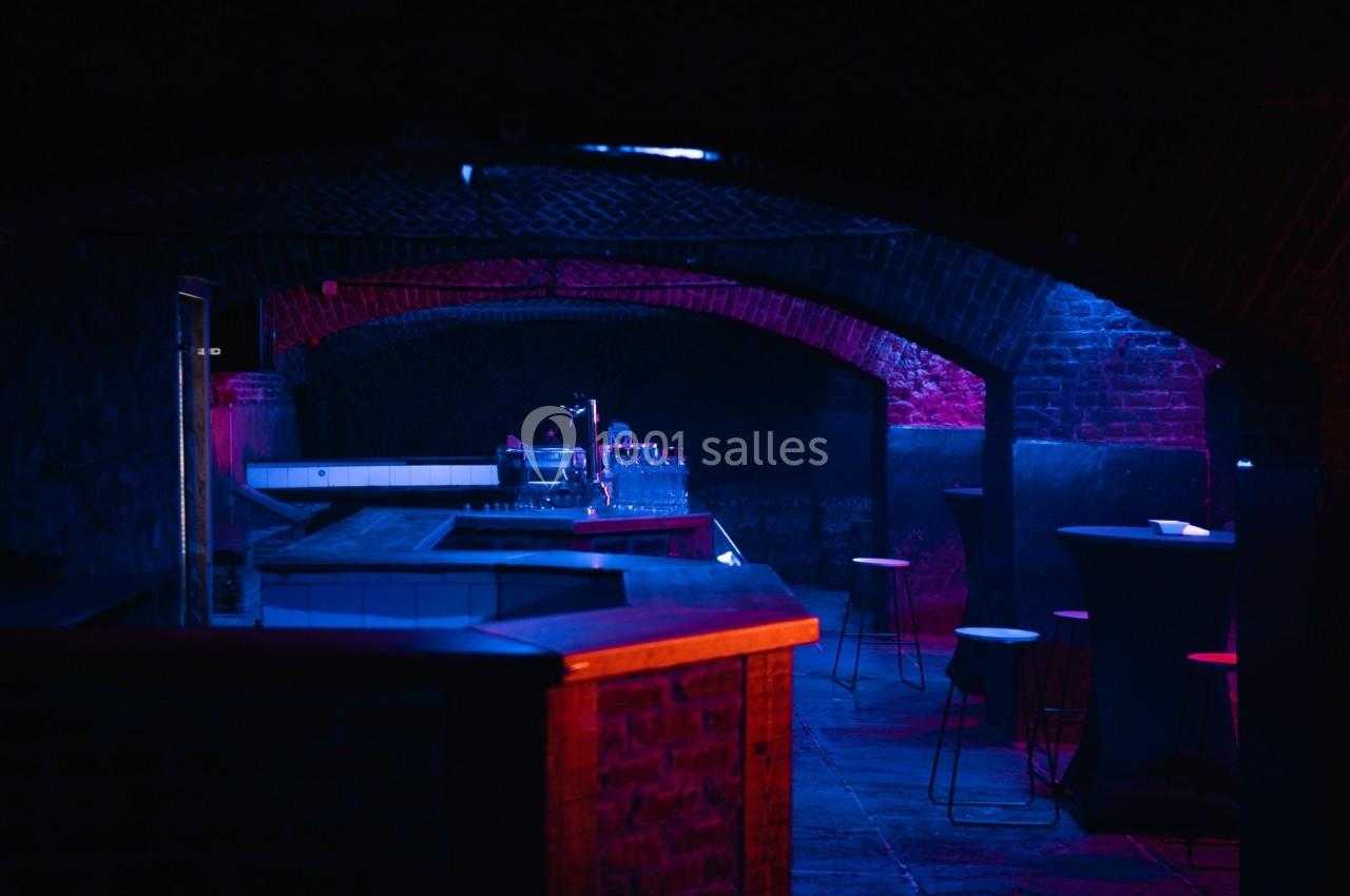 Bar souterrain avec éclairage tamisé bleu et rose, comptoirs en bois et tabourets dispersés.