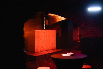 Salle sombre avec murs en briques, éclairage tamisé, scène en bois et tables basses au premier plan.