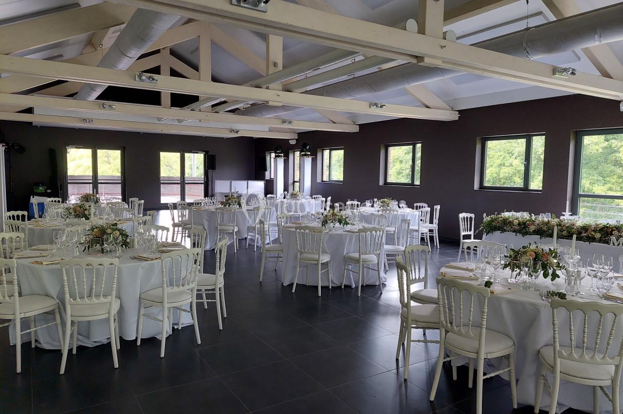 Salle de réception lumineuse avec tables rondes dressées, chaises blanches et poutres apparentes au plafond.