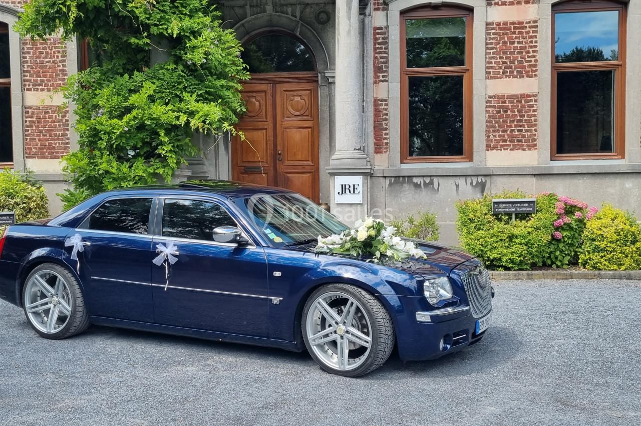 Voiture de mariage bleue décorée de fleurs blanches, garée devant un bâtiment en briques avec porte en bois.