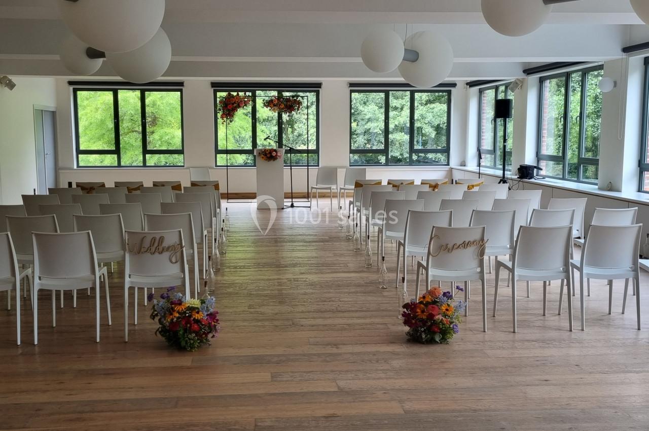 Salle lumineuse aménagée pour une cérémonie, avec des chaises blanches alignées et des bouquets de fleurs colorées.