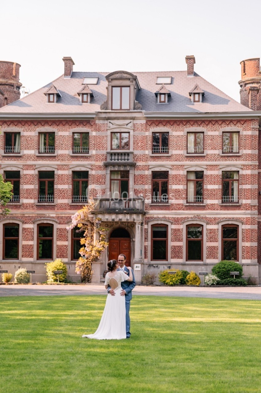Un couple en tenue de mariage se tient devant un grand bâtiment en briques rouges entouré de verdure.