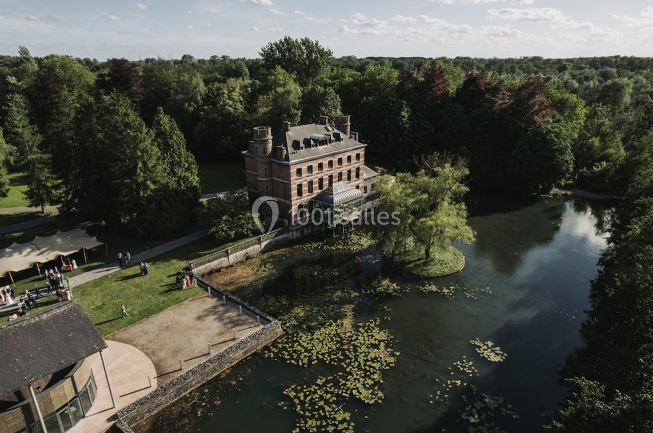 Vue aérienne d'un manoir entouré d'arbres, bordé par un étang avec des nénuphars et une pelouse aménagée.